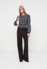 Navy en witte geruite blouse met pofmouwen, gecombineerd met hooggesneden donkere broeken. Houdt een kleine witte handtas vast. Gladde stof textuur.