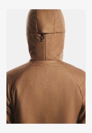 Person, die eine braune gepolsterte Jacke mit Kapuze trägt, von hinten vor einem schlichten, hellen Hintergrund gezeigt.