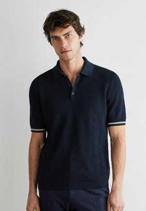 Junger Mann mit zerzaustem braunem Haar trägt ein dunkelblaues kurzärmeliges Strick-Polohemd mit weißer Einfassung an den Ärmeln und steht vor einer schlichten hellen Wand.