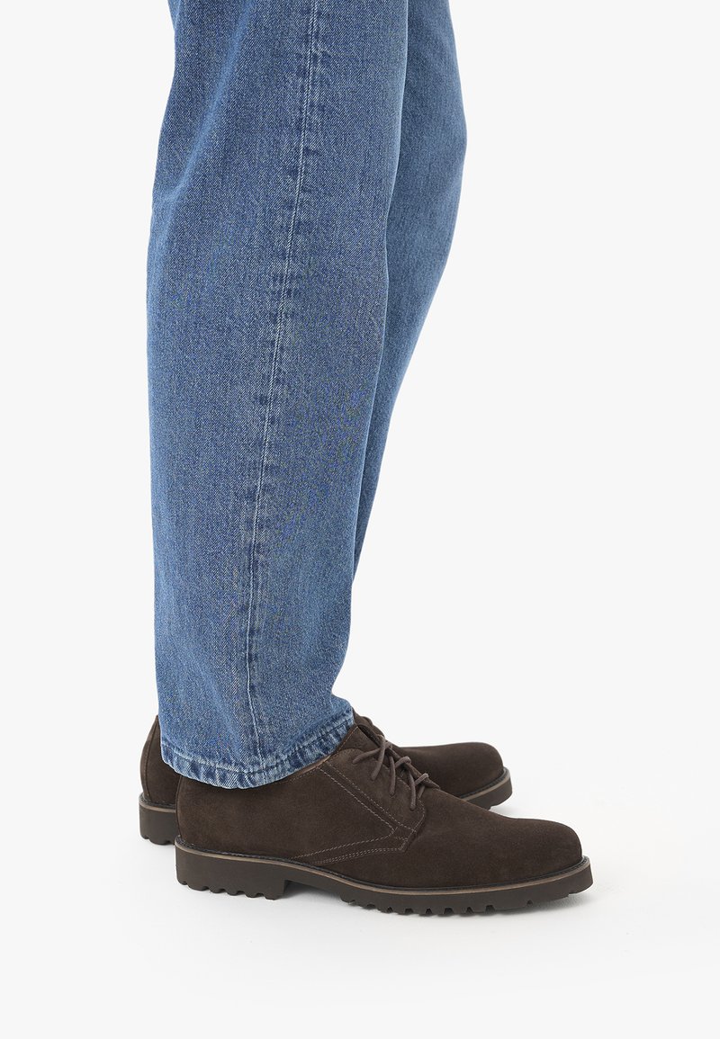 Personne portant des chaussures en daim marron à lacets et un jean bleu, debout sur une surface blanche, vue de profil.