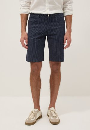 Mann trägt marineblaue knielange Shorts mit Nadelstreifen, weißes Hemd mit langen Ärmeln und beige Turnschuhe, steht vor einfachem Hintergrund.