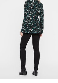 Blusa nera a fantasia con forme astratte nelle tonalità verde teal e beige, vita aderente, maniche lunghe, abbinata a jeans skinny neri e stivali con tacco a blocco.