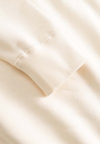 Tissu de sweat-shirt beige clair en gros plan, avec un poignet côtelé texturé, une surface lisse, et un design simple sans motifs ni éléments métalliques.