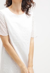 Valkoinen mekko, jossa on yksityiskohtainen kirjailu pyöreillä kuvioilla, lyhyet pitsihihat ja pyöreä pääntie. Sileä, kevyt kangas.