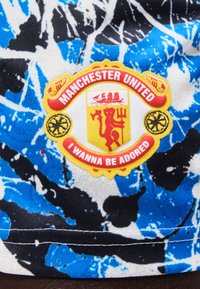 Σήμα της Manchester United με κίτρινη ασπίδα, κόκκινο διάβολο που κρατά τρίαινα, καράβια και το κείμενο "I WANNA BE ADORED" σε μπλε, λευκό και μαύρο υφαντό ύφασμα.