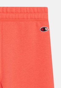 Coral sweatpants with an elastic waistband. Features the Champion logo embroidered in blue and red on the left side. Soft texture, solid color.