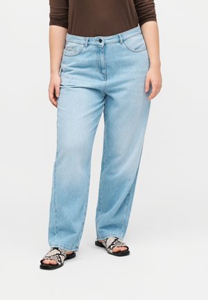 Oseba, oblečena v svetlo modre ravne kavbojke, rjavo dolg rokav majico in črno-bele sandale z vzorcem kačje kože, stoji na belem ozadju.