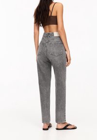 Femme aux cheveux longs portant un jean en denim gris taille haute, un crop top marron et des sandales noires, debout devant un fond blanc.