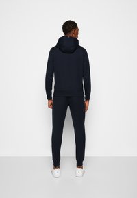 Felpa con cappuccio blu navy abbinata a pantaloni della tuta, dotata di cappuccio con cordino, polsini a costine e sneakers bianche. Design semplice e monocromatico.