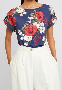 Top floreale blu navy con grandi rose rosse e fiori bianchi, realizzato in tessuto leggero con una vestibilità ampia e maniche corte arrotolate.