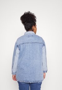 Kobieta z kręconymi włosami, związanymi w wysoki kok, ubrana w jasnoniebieską dżinsową kurtkę i dżinsy, stojąca przodem do gładkiego białego tła.