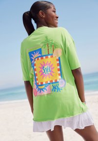Maglietta oversize verde brillante con un design grafico colorato che presenta palme, soli e conchiglie sul retro. Materiale in cotone.