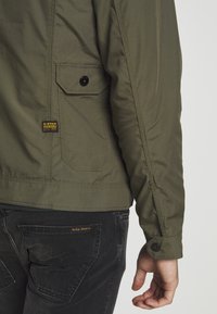 Olivgrön jacka med fickdetalj på vänster sida, med svarta knappstängningar och en sydd fåll. Bärs med mörka jeans.