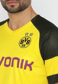 Gul och svart sporttröja med V-ringning, som har ett texturerat svart axelområde, klubbens logotyp och "EVONIK" i fet lila text.