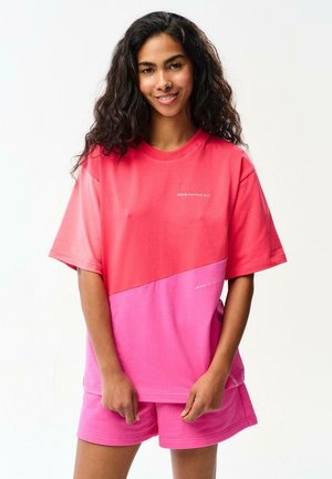 Uśmiechnięta kobieta z długimi falującymi włosami ma na sobie różowy t-shirt w stylu color-block oraz pasujące szorty, stojąc na tle jasnego, jednolitego tła.