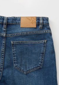 Jeans in denim con lavaggio blu, dotati di una toppa in pelle testurizzata in vita e una tasca posteriore singola con dettagli di cucitura visibili.