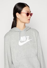 Grijze hoodie van zacht materiaal met een groot wit Nike-logo. Draagt het met doorschijnende, roze cat-eye zonnebril.