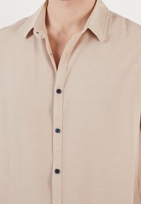 Camisa beige de manga corta en tela texturizada con un ligero efecto arrugado. Cuenta con cuello y botones azul oscuro en la tapeta frontal.