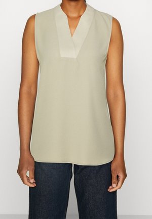 Ærmeløs beige bluse med en bred V-hals krave båret med mørkeblå jeans, vist fra skuldre til hofter.