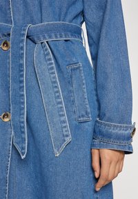 Közékkék farmer trench coat. Jellemzője egy megkötős derékövr, gombos zárás és varrásmintás mandzsetta. Kiegészítők között található egy foltujj zseb.