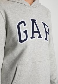 Sweat à capuche gris en mélange de coton, avec un grand logo "GAP" brodé en bleu marine avec un contour blanc. Comprend une poche avant.