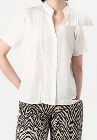 Osoba v bílé krátké košili na knoflíky s krajkovými detaily a béžových kalhotách s černými zebřími pruhy, stojící s rukama v kapsách.