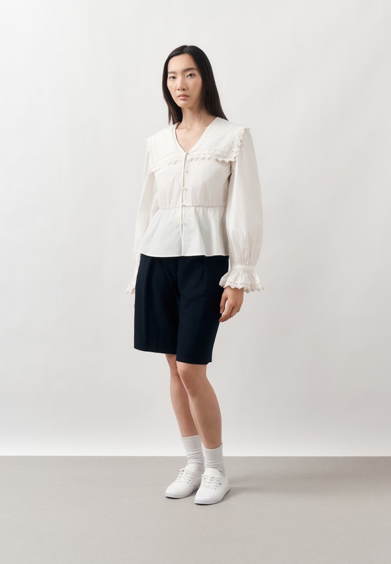 Femme debout portant un chemisier blanc avec col et poignets en dentelle, un short noir aux genoux, des chaussettes blanches et des baskets blanches sur un fond neutre.