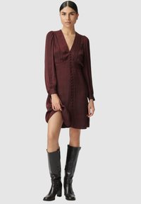 Femme portant une robe boutonnée à manches longues bordeaux avec des plis, tenant l'ourlet, associée à des bottes en cuir noires jusqu'aux genoux.