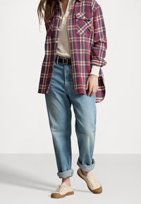 Person, der ein rot-blau kariertes Hemd, ein cremefarbenes Henley-Oberteil, lockere hellblaue Jeans mit Umschlägen, einen schwarzen Gürtel und beige Sneaker mit Gummisohlen trägt.