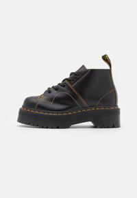 Botas de tobillo de cuero negro con costuras amarillas, suela de goma gruesa y diseño de puntera redonda. Presenta una lengüeta en el talón.