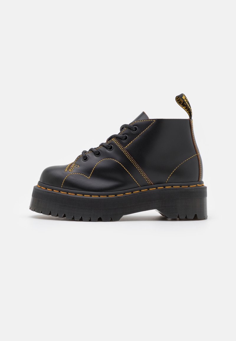Botas de tobillo de cuero negro con costuras amarillas, suela de goma gruesa y diseño de puntera redonda. Presenta una lengüeta en el talón.