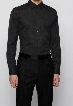 Mężczyzna ubrany w czarną, zapinaną na guziki koszulę z długim rękawem oraz pasujące czarne spodnie z subtelnym teksturowanym wzorem na jednolitym tle.