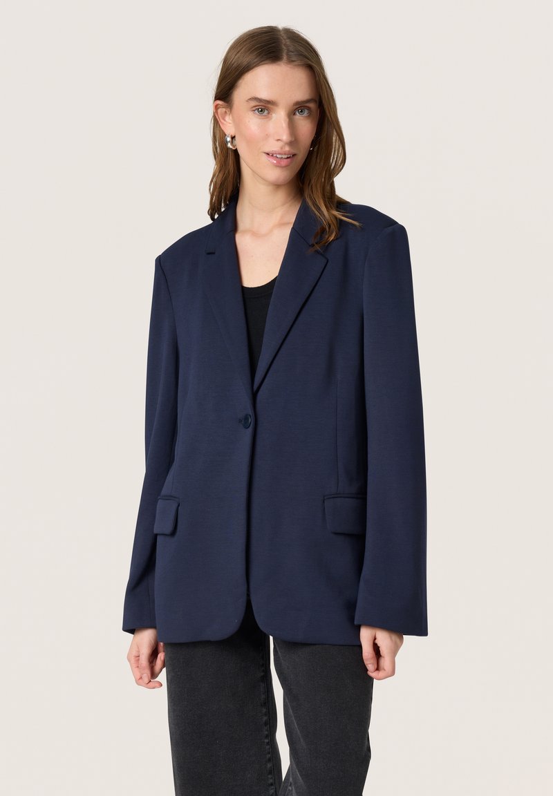 Marineblå skræddersyet blazer med en enkelt knap, to forreste lommer og en glat tekstur. Båret over en sort top og parret med mørke jeans.
