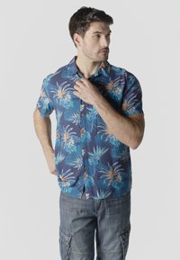 Hombre ajustando el cuello de una camisa hawaiana azul de manga corta con patrón floral, vestido con pantalones cortos de mezclilla tipo cargo, de pie frente a un fondo liso.