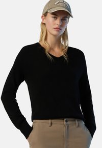 Eine Frau trägt einen schwarzen V-Ausschnitt-Pullover, beige Hosen mit den Händen in den Taschen und eine beigefarbene Mütze mit weißem Text. Sie steht vor einem schlichten Hintergrund.