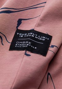 Tessuto rosa con un'etichetta nera che recita "IL TUO UNICO LIMITE È LA TUA MENTE OMBRE STREETSTYLE dal 1985." Presenta linee scure curve sullo sfondo.