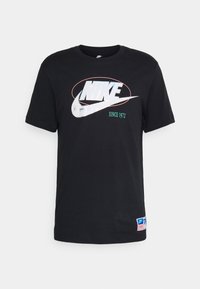 Schwarzes Baumwoll-T-Shirt mit einem großen weißen Nike-Logo, einem silbernen Swoosh und grüner Schrift, sowie einem farbigen Etikett auf der unteren linken Seite.