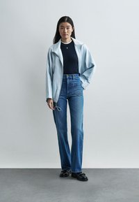 Νέα γυναίκα στέκεται φορώντας ανοιχτό μπλε μπουφάν, μαύρο ριμπ μπλουζάκι, ψηλόμεσα μπλε τζιν και μαύρες μοκασίνες μπροστά σε απλό γκρι φόντο.