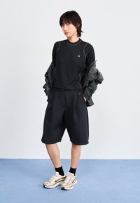 Black long-sleeve top with gray accents, paired with loose black shorts. Model wears gold and white athletic shoes. Plain background.