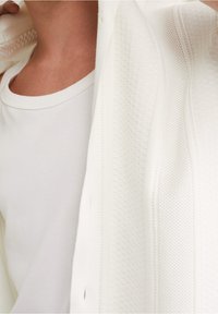 Vit stickad kofta med texturerade mönster och knappdetaljer, lagd över en enkel vit t-shirt med rund hals. Mjukt tyg, avslappnad design.