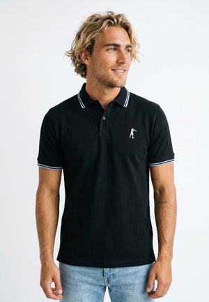 Junger Mann mit blonden Haaren, der ein schwarzes Poloshirt mit weißem Saum und einer kleinen weißen bestickten Figur trägt, kombiniert mit hellblauen Jeans.