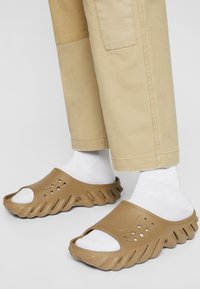 Brown plastic slide sandals with an open toe design, featuring ventilation holes and a textured sole, worn with white ankle socks.