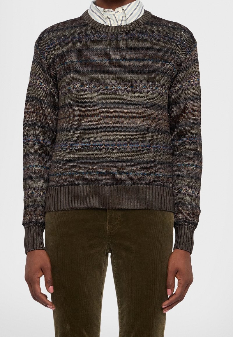 Homme portant un pull en maille rayé foncé par-dessus une chemise à rayures blanches et bleues avec col et un pantalon en velours côtelé vert olive.