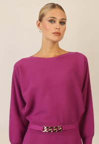 Suéter de punto ribeteado fucsia con escote amplio y mangas largas, que presenta un cinturón de cadena dorada en la cintura. Tejido suave y texturizado.