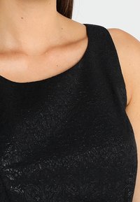 Czarna sukienka z teksturowanym, skomplikowanym wzorem. Posiada szeroki, zaokrąglony dekolt i bezrękawowy krój, podkreślający blask materiału.