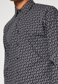 Camisa negra de botones con un patrón repetitivo de escritura blanca. Cuello clásico y detalle de un solo bolsillo en el lado izquierdo.