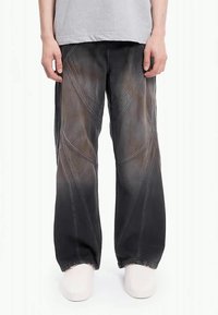 Oseba nosi zbledela črna široka hlače z ukrivljenimi šivi, v kombinaciji z svetlo sivo majico in belimi čevlji, stoji ob belem ozadju.