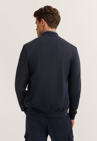 Mężczyzna z krótkimi brązowymi włosami, ubrany w ciemnoniebieską długą kurtkę z długim rękawem oraz ciemne spłaszczone spodnie, stojący plecami do obiektywu na jednolitym tle.