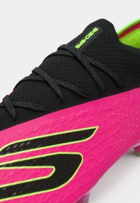 Musta ja vaaleanpunainen jalkapallokengä, jossa on teksturoitu pinta, vihreät yksityiskohdat ja erottuva musta logo. Varustettu vahvoilla nauhoilla ja matalalla leikkauksella.