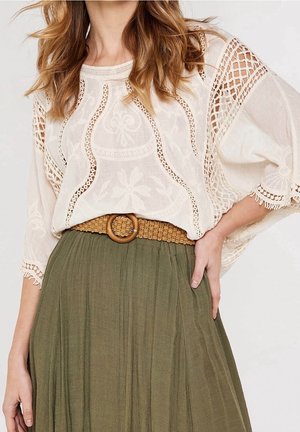 Femme portant un chemisier blanc à détails en dentelle, rentré dans une jupe plissée taille haute vert olive avec une ceinture marron tissée.